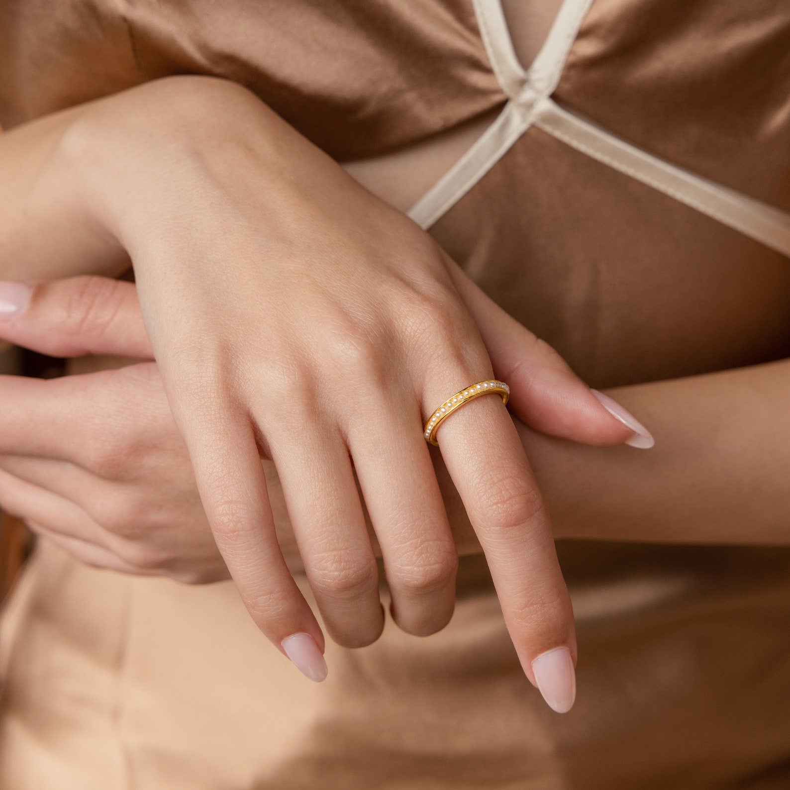 Close-up of a woman's hand wearing the Alicia Pearl Inlay Band, a gold stackable ring with pearl details, against a beige satin dress.