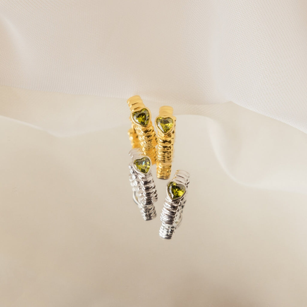 The Peridot Heart Huggies set includes two pairs of minimalistic hoop earrings—one gold, one silver—each accented with a vibrant green peridot heart gem for a subtle pop of color.