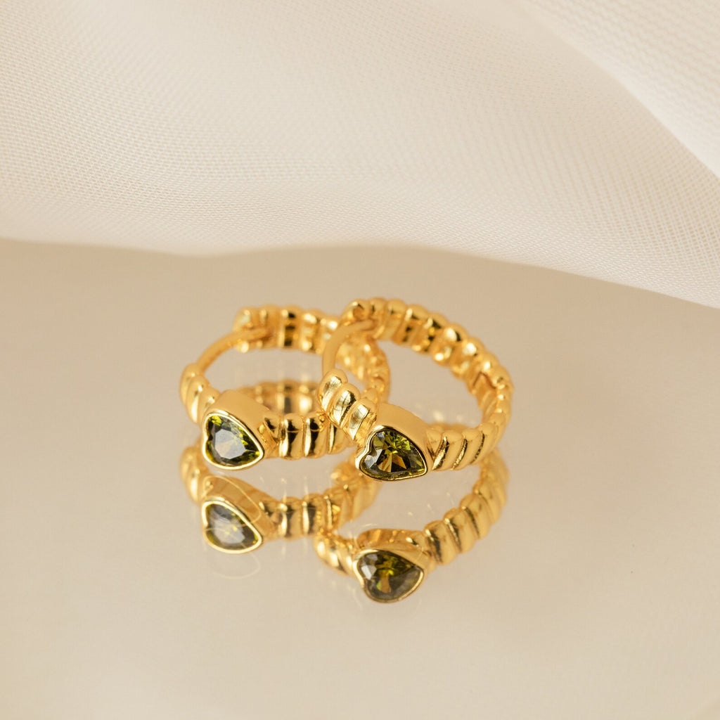 The Peridot Heart Huggies, two gold rings with green gemstone settings, are displayed on a reflective surface with a white fabric backdrop, evoking the look of minimalistic earrings.