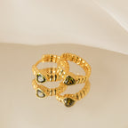 The Peridot Heart Huggies, two gold rings with green gemstone settings, are displayed on a reflective surface with a white fabric backdrop, evoking the look of minimalistic earrings.