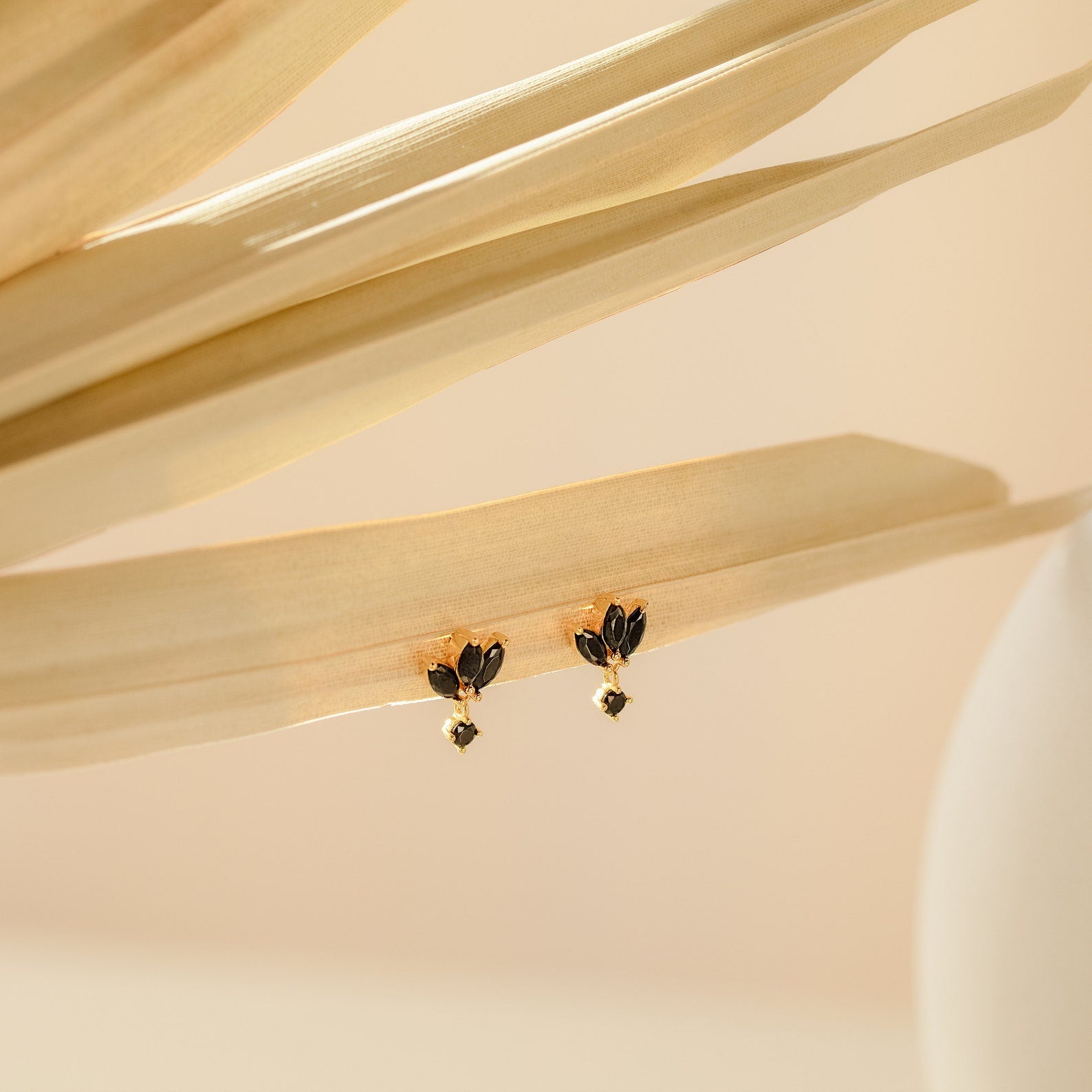 The Emily Drop Earrings in gold and black rest on dried beige palm leaves, set against a minimal, soft background—ideal statement earrings for adding a subtle hint of vintage elegance.