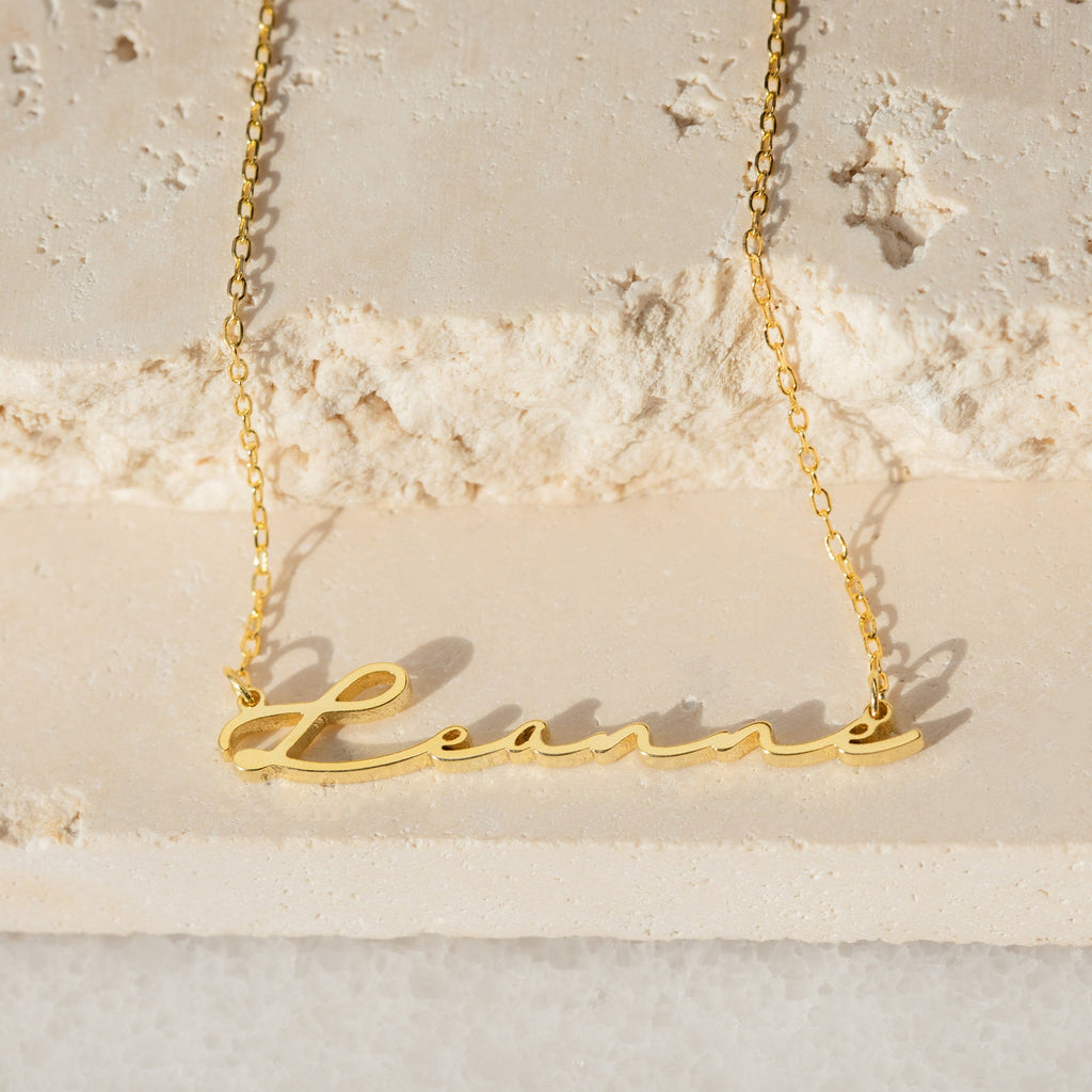 Minimal Sideways Name Necklace in gold with the name "Leane" in cursive, featuring an off-center design and displayed on a textured stone surface.