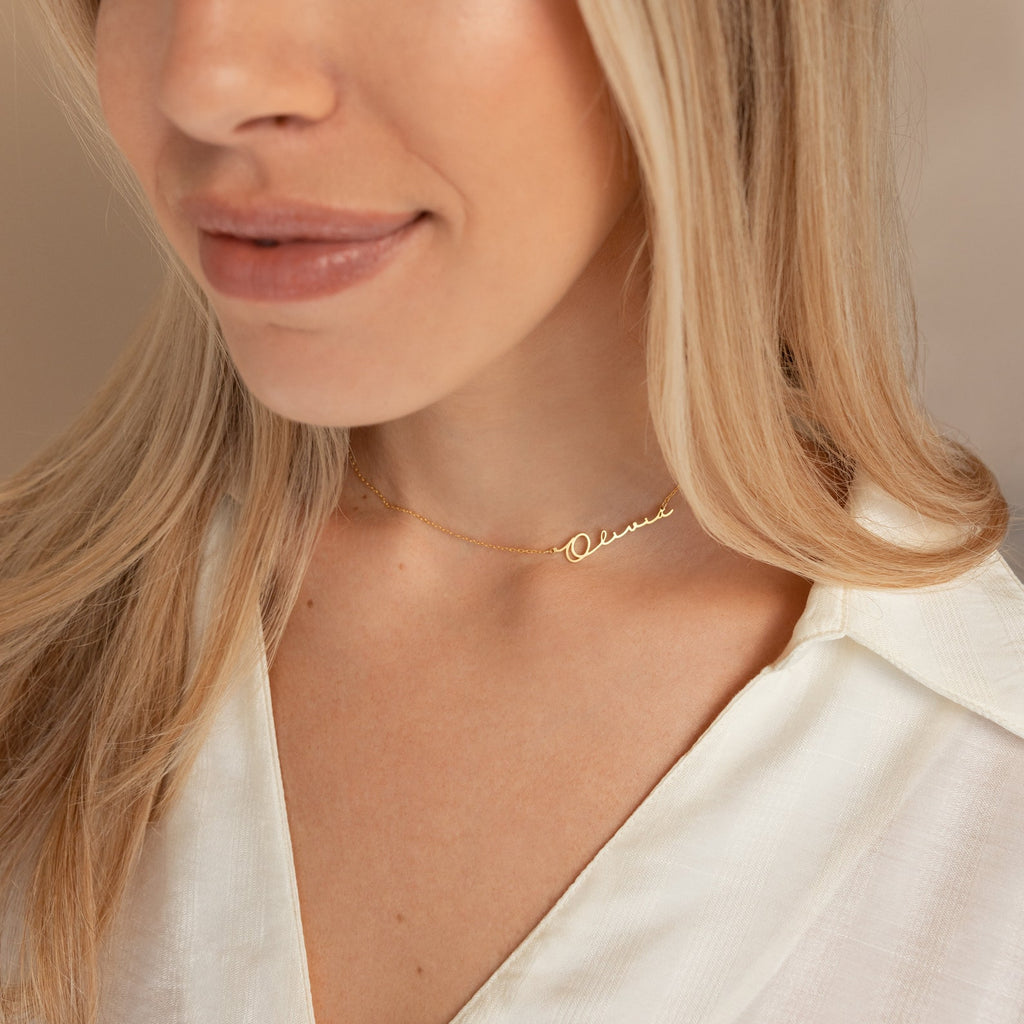 A woman wears a white shirt and the Minimal Sideways Name Necklace, featuring an off-center gold name pendant; only her lower face and neck are shown.