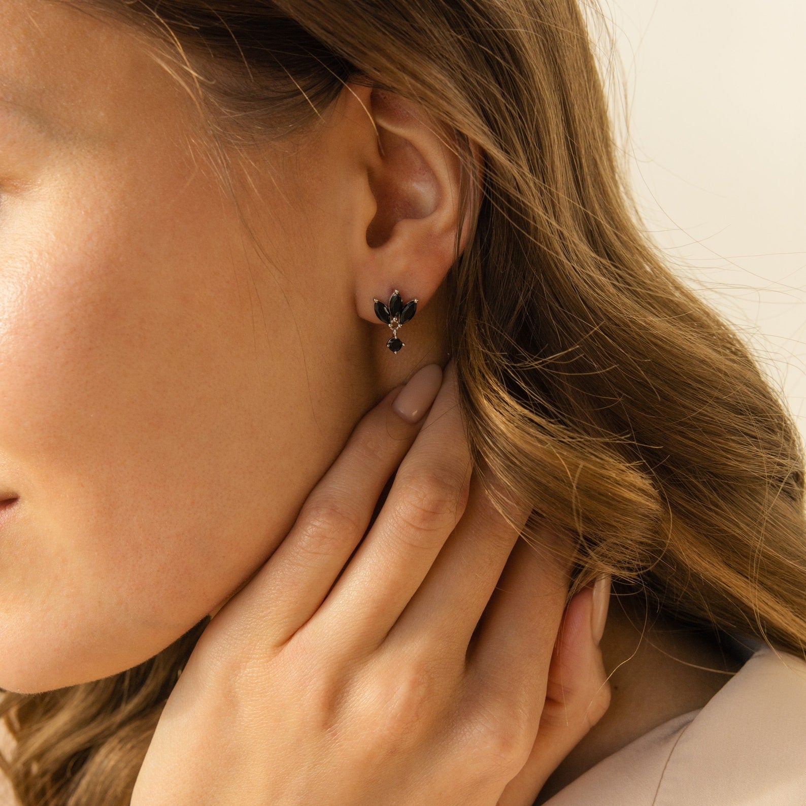A woman with wavy hair wears delicate Emily Drop Earrings, shaped like black petal birds, and gently touches her ear.