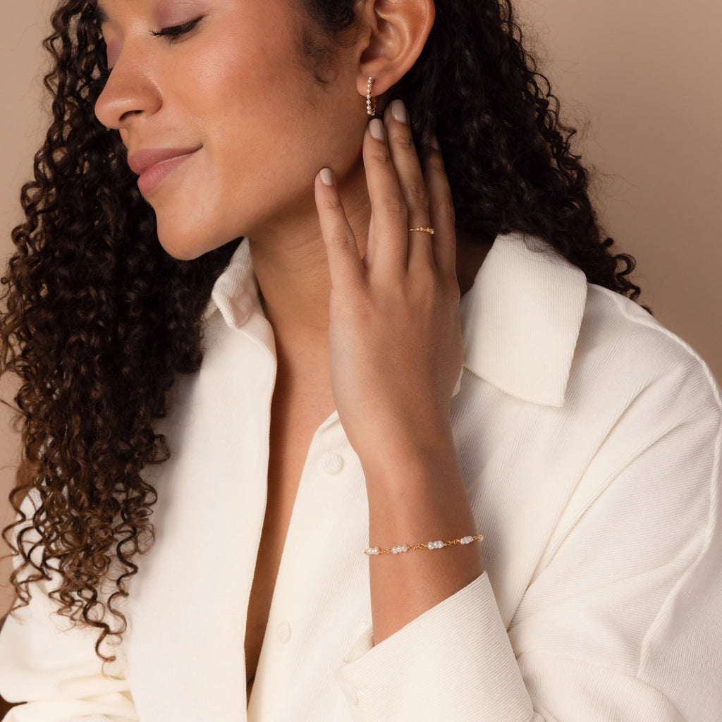 Woman with curly hair wearing gold jewelry and the Hali Pearl Station Bracelet, dressed in a cream shirt against a beige background—an elegant choice for bridal jewelry or the perfect bridesmaid gift.