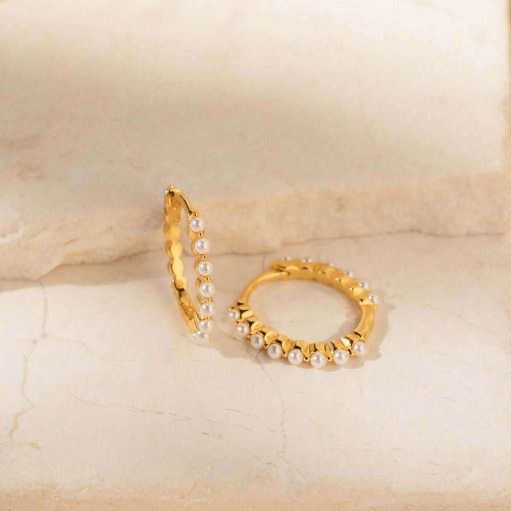 The Malik Pearl Hoops feature gold hoops accented with small white pearls, shown on a light stone surface. These minimalist earrings are perfect as wedding jewelry or for adding elegance to everyday outfits.