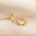 The Malik Pearl Hoops feature gold hoops accented with small white pearls, shown on a light stone surface. These minimalist earrings are perfect as wedding jewelry or for adding elegance to everyday outfits.