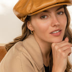 Woman in a tan cap and jacket, smiling with hand resting on chin, wearing elegant minimalist earrings—the Malik Pearl Hoops—and a gold hoop earring.