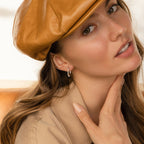 Woman in a tan outfit and brown cap, touching her neck while wearing Malik Pearl Hoops, looking at the camera.