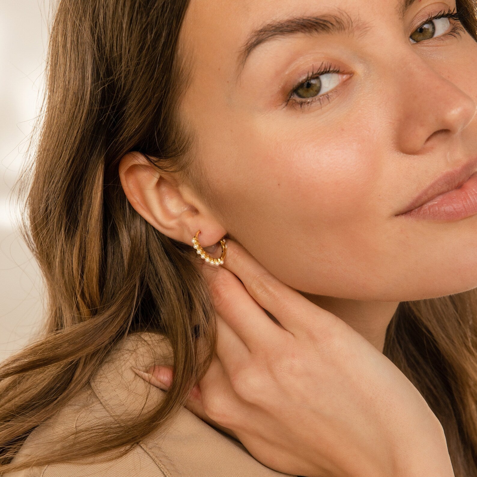A woman with light brown hair smiles softly at the camera, touching her minimalist Malik Pearl Hoops earring.