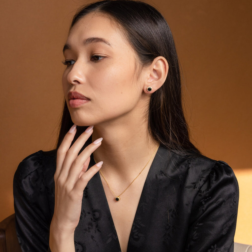 A woman in a black outfit wears matching gold jewelry with black stones, featuring the striking Halo Birthstone Necklace, as she looks to the side against a brown background.