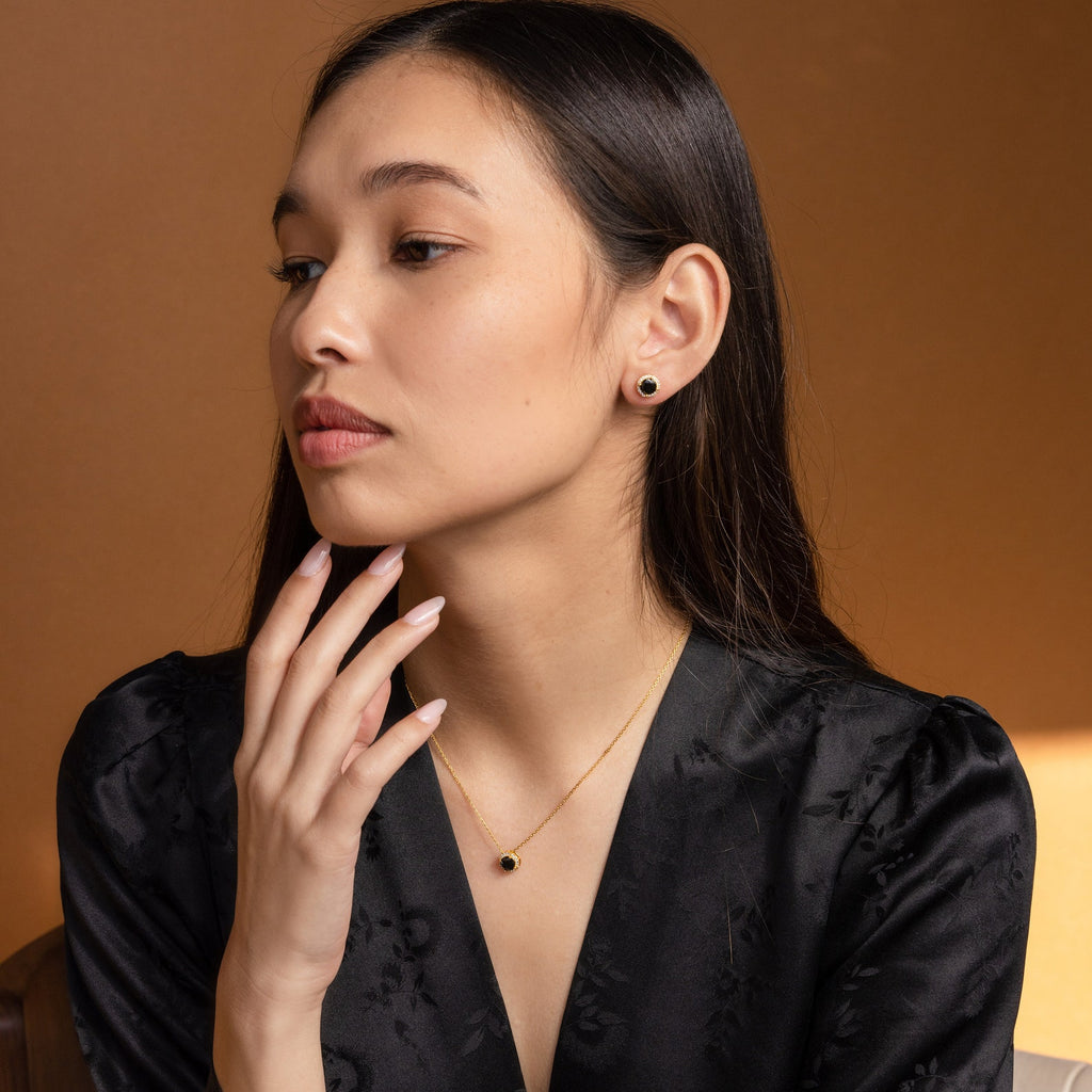 Woman in a black dress wears Halo Birthstone Studs—gold earrings with black stones—in a vintage style, looking to the side against a brown background.