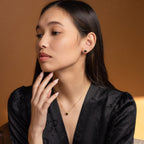 Woman in a black dress wears Halo Birthstone Studs—gold earrings with black stones—in a vintage style, looking to the side against a brown background.