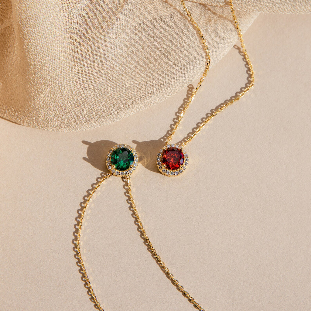 Two Halo Birthstone Necklaces featuring round green and red gemstone pendants are displayed on a beige fabric and surface.