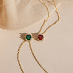 Two Halo Birthstone Necklaces featuring round green and red gemstone pendants are displayed on a beige fabric and surface.