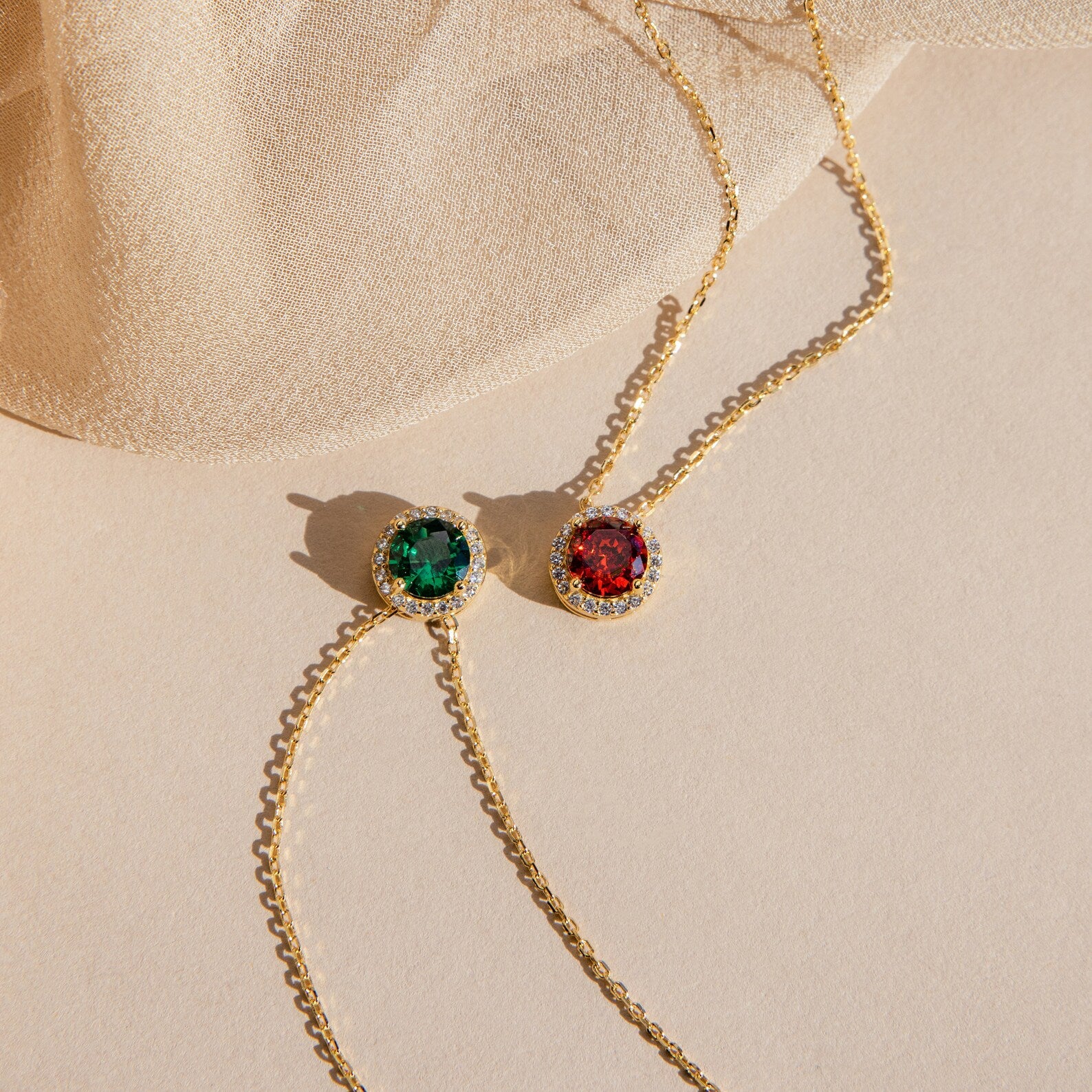 Two Halo Birthstone Necklaces featuring round green and red gemstone pendants are displayed on a beige fabric and surface.