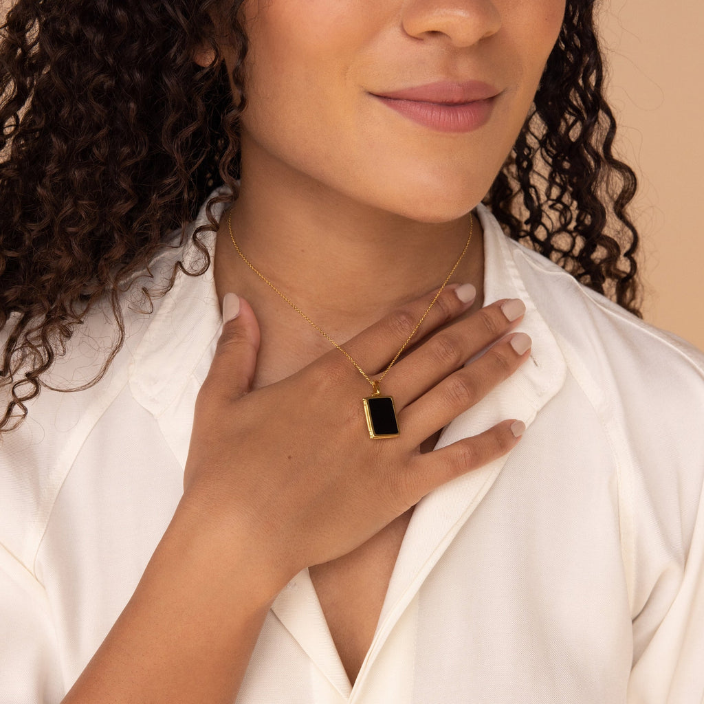 A woman wears a white blouse and the Onyx Locket Necklace—a gold chain with a black enamel pendant—her hand resting on her chest.
