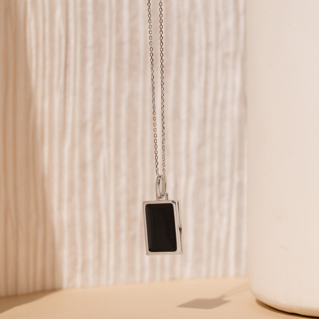 The Onyx Locket Necklace features a sleek rectangular black enamel pendant on a silver chain, set against a soft beige textured background—an elegant and personalized gift choice.