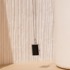 The Onyx Locket Necklace features a sleek rectangular black enamel pendant on a silver chain, set against a soft beige textured background—an elegant and personalized gift choice.
