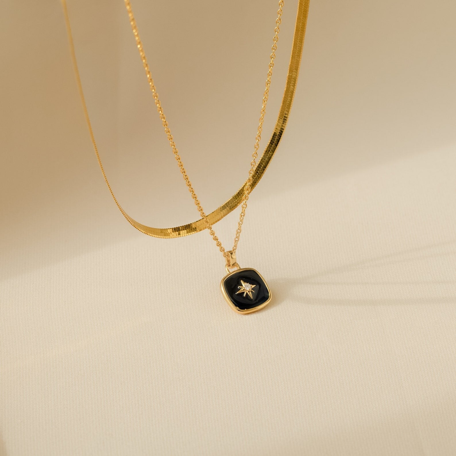 The Hermoine Pendant Necklace and another gold necklace, featuring a black and gold star pendant with Mother of Pearl, are elegantly displayed on a beige background.