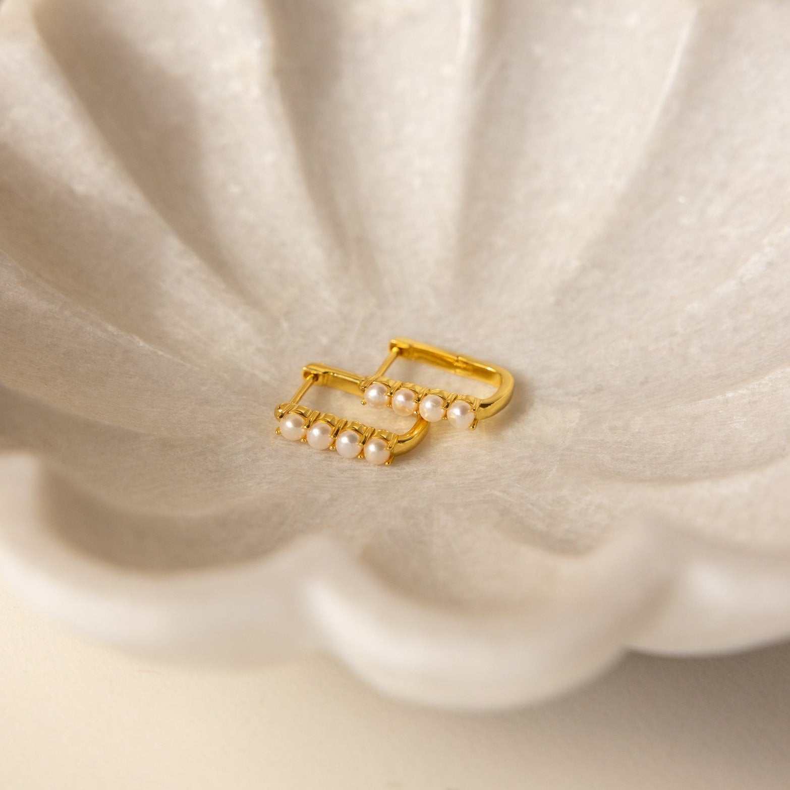 The Rayne Pearl Link Earrings, featuring gold hoops with small freshwater pearls, rest in a white scalloped ceramic dish—an elegant minimalist jewelry accent.