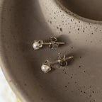 The Raindrop Pearl Onyx Studs feature pearl and black onyx accents with silver backs, elegantly arranged on a textured, round gray dish.