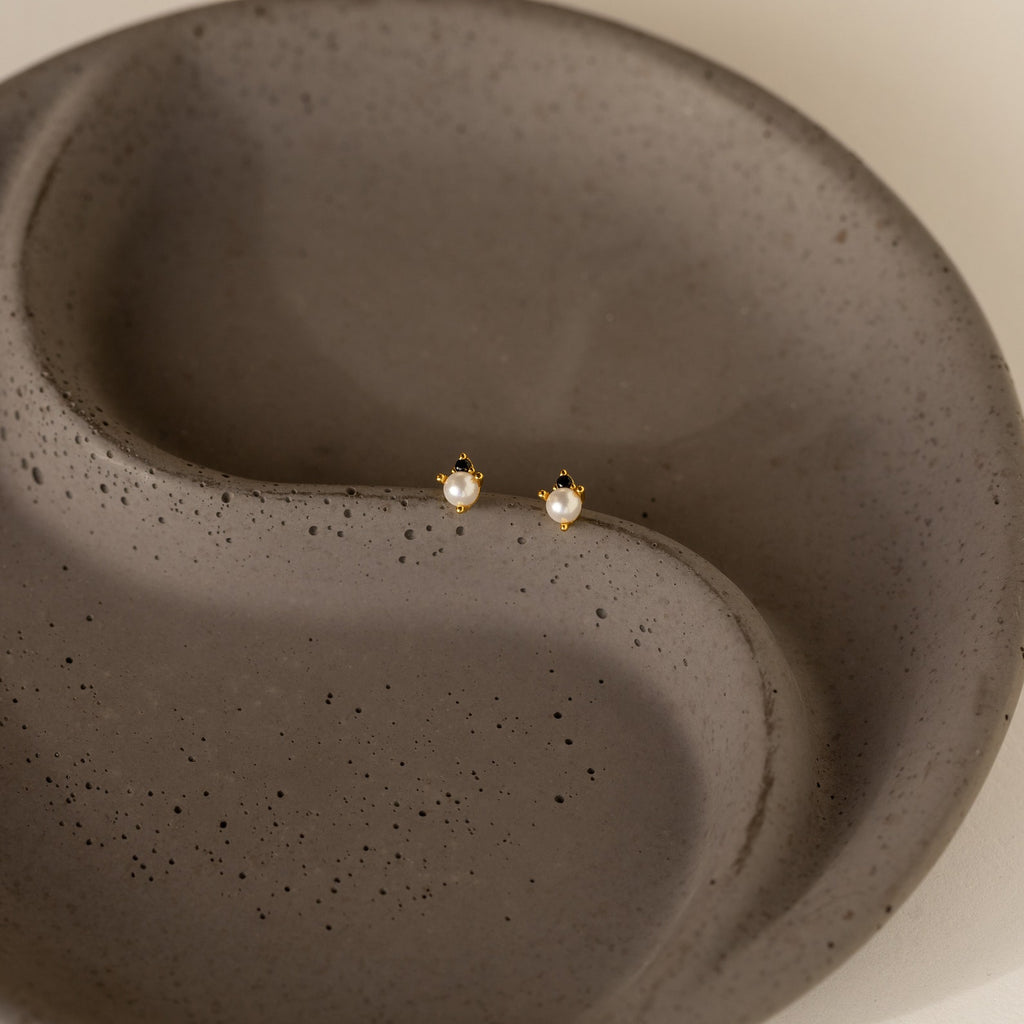 Raindrop Pearl Onyx Studs, gold earrings featuring small pearls, evoke classic pearl studs and are displayed on a round, textured gray ceramic dish.
