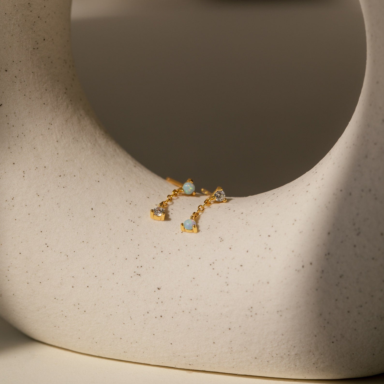 Opal Drop Studs with delicate chains and blue and clear gemstones, showcased on a smooth, speckled ceramic surface.