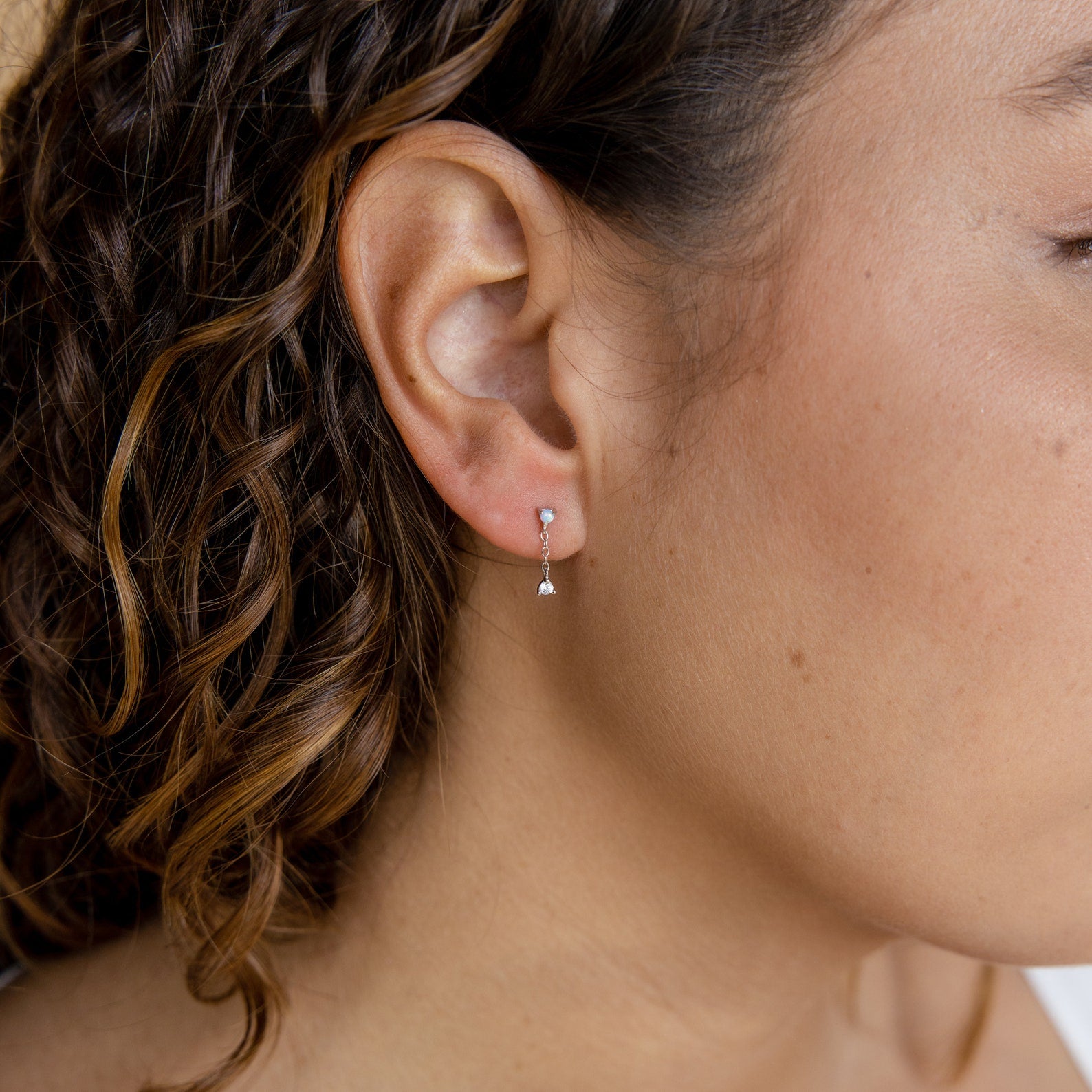 Close-up of a woman's ear wearing Opal Drop Studs—small silver hoops with a single dangling opal gemstone, curly hair framing the look.