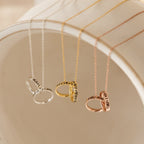 The Engraved Interlocking Necklace features three personalized name rings in silver, gold, and rose gold, displayed on a white curved surface—an ideal anniversary gift with custom engraving.