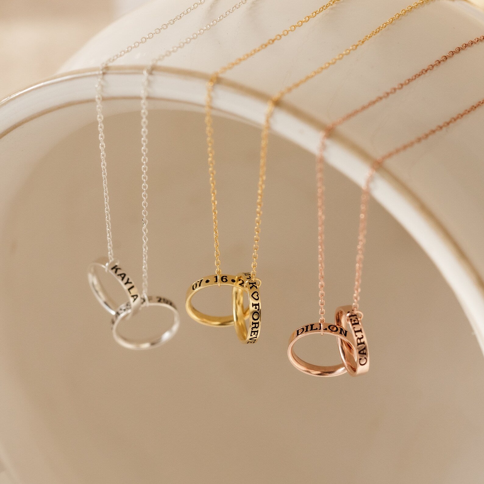 The Engraved Interlocking Necklace features three personalized name rings in silver, gold, and rose gold, displayed on a white curved surface—an ideal anniversary gift with custom engraving.