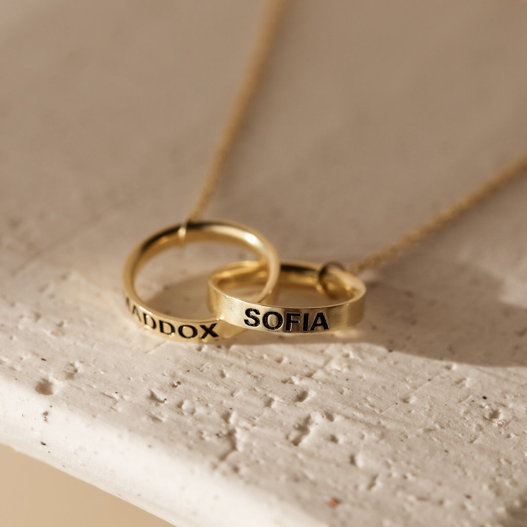 The Engraved Interlocking Necklace features two gold rings personalized with MADDOX and SOFIA, elegantly intertwined on a delicate gold chain against a light background.