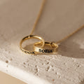 The Engraved Interlocking Necklace features two gold rings personalized with MADDOX and SOFIA, elegantly intertwined on a delicate gold chain against a light background.