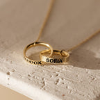 The Engraved Interlocking Necklace features two gold rings personalized with MADDOX and SOFIA, elegantly intertwined on a delicate gold chain against a light background.