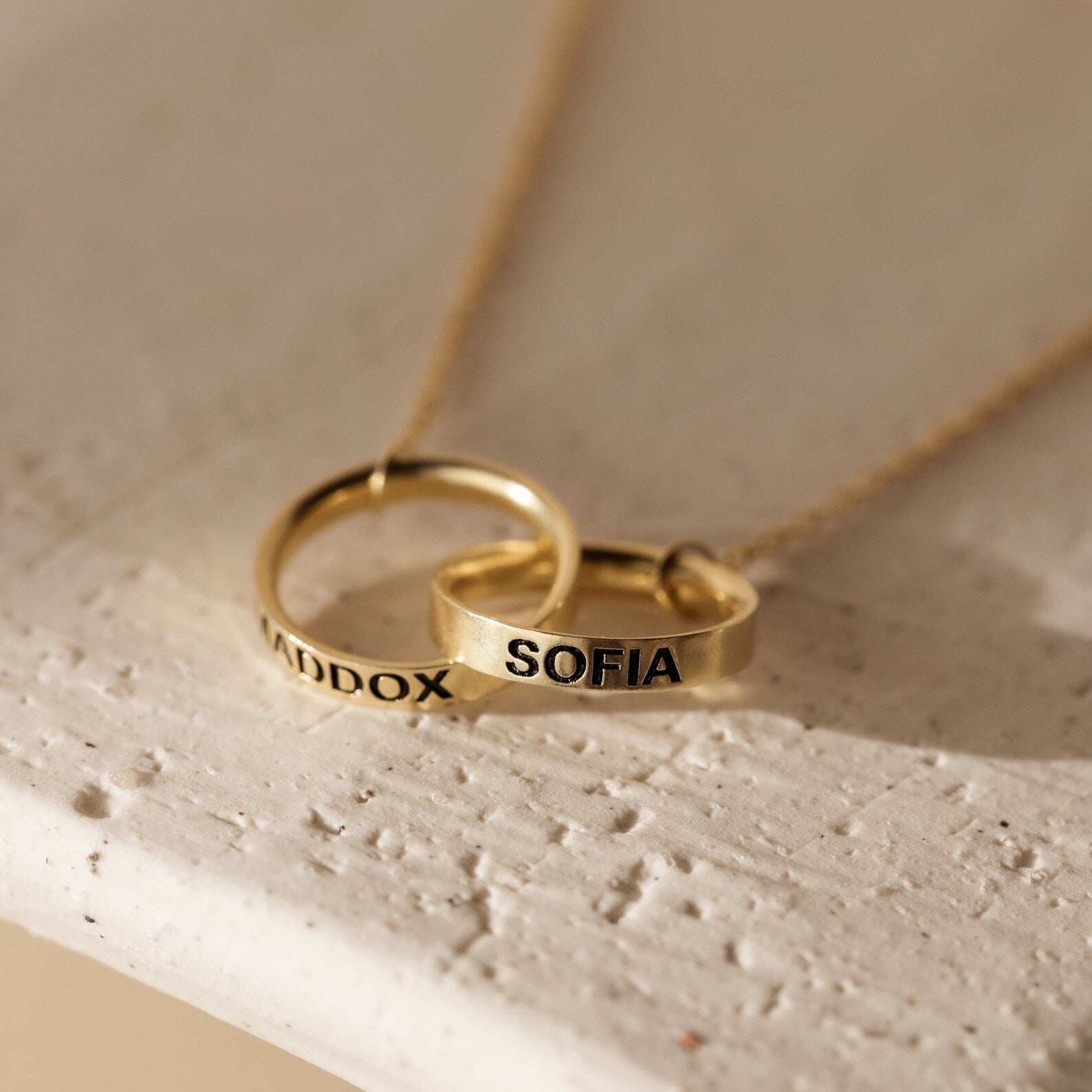 The Engraved Interlocking Necklace features two gold rings personalized with MADDOX and SOFIA, elegantly intertwined on a delicate gold chain against a light background.