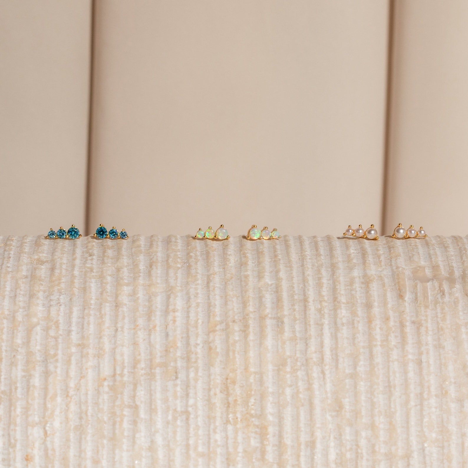 The Triple Birthstone Studs—blue, green, and pearl pairs—are displayed on a textured beige surface, making them a thoughtful gift for mothers, daughters, or anyone who loves elegant birthstone earrings.