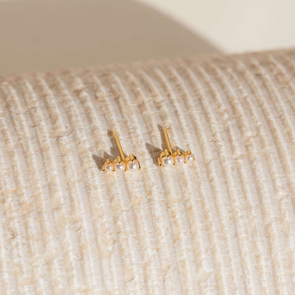 Triple Birthstone Studs featuring gold and small pearls, reminiscent of elegant birthstone earrings, displayed on a textured, cream-colored surface.