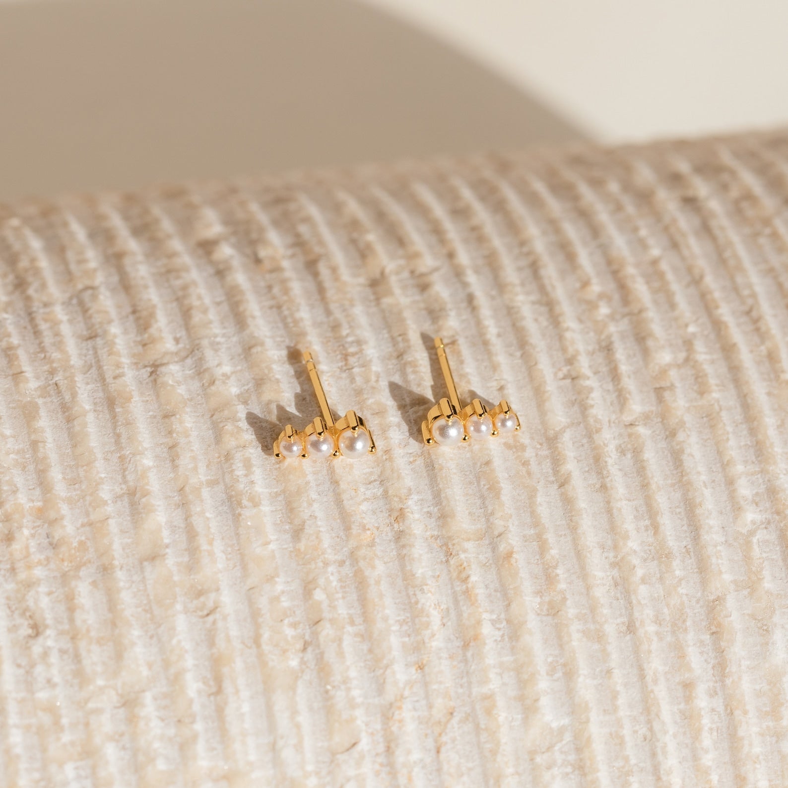 Triple Birthstone Studs featuring gold and small pearls, reminiscent of elegant birthstone earrings, displayed on a textured, cream-colored surface.