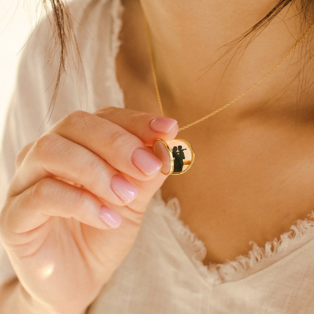 A woman wearing a cream top holds a meaningful gift—a Pearl Disc Locket Necklace featuring a gold locket with a black cat silhouette inside.