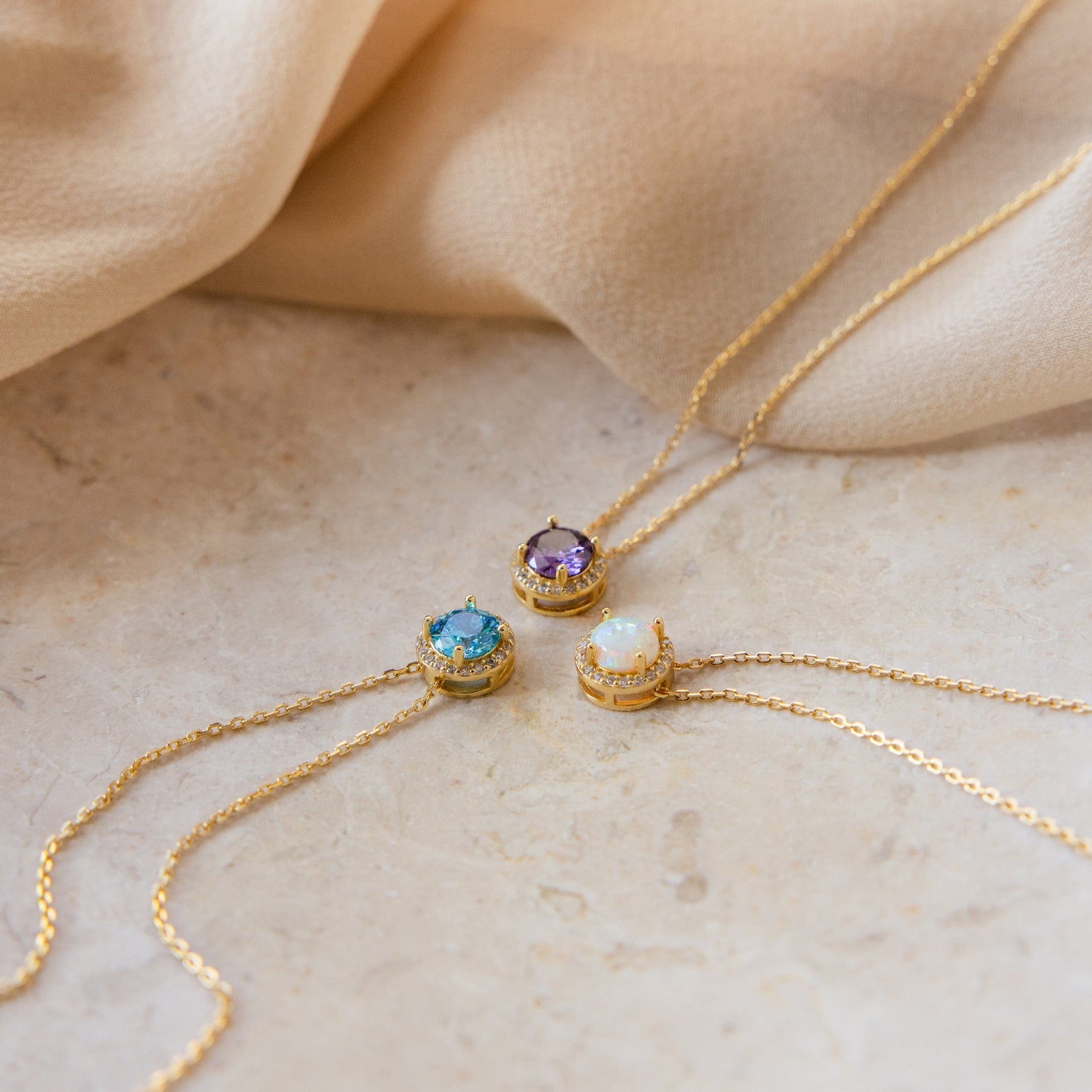 Three Halo Birthstone Necklaces—featuring purple, blue, and opal gemstones—are displayed on beige fabric and stone, each providing a modern take on the classic birthstone necklace.