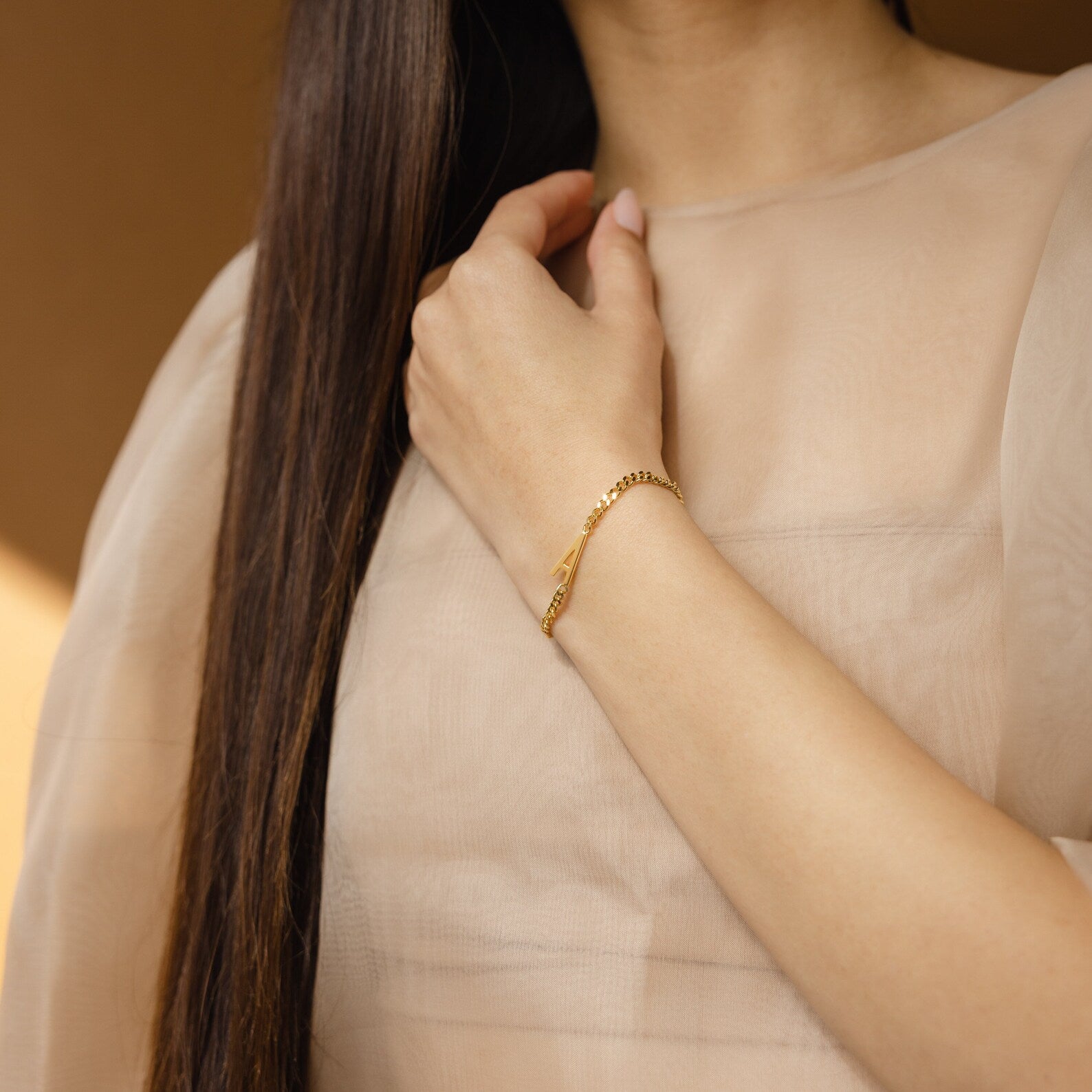 A woman in a sheer beige top, with long brown hair over her shoulder, wears the Large Letter Bracelet—an elegant piece perfect for everyday jewelry.