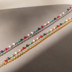 The Rainbow Station Bracelet features double chains—one in silver, one in gold—with vibrant colorful beads. Handmade and displayed on a neutral surface, it's perfect for adding a playful pop to any look.