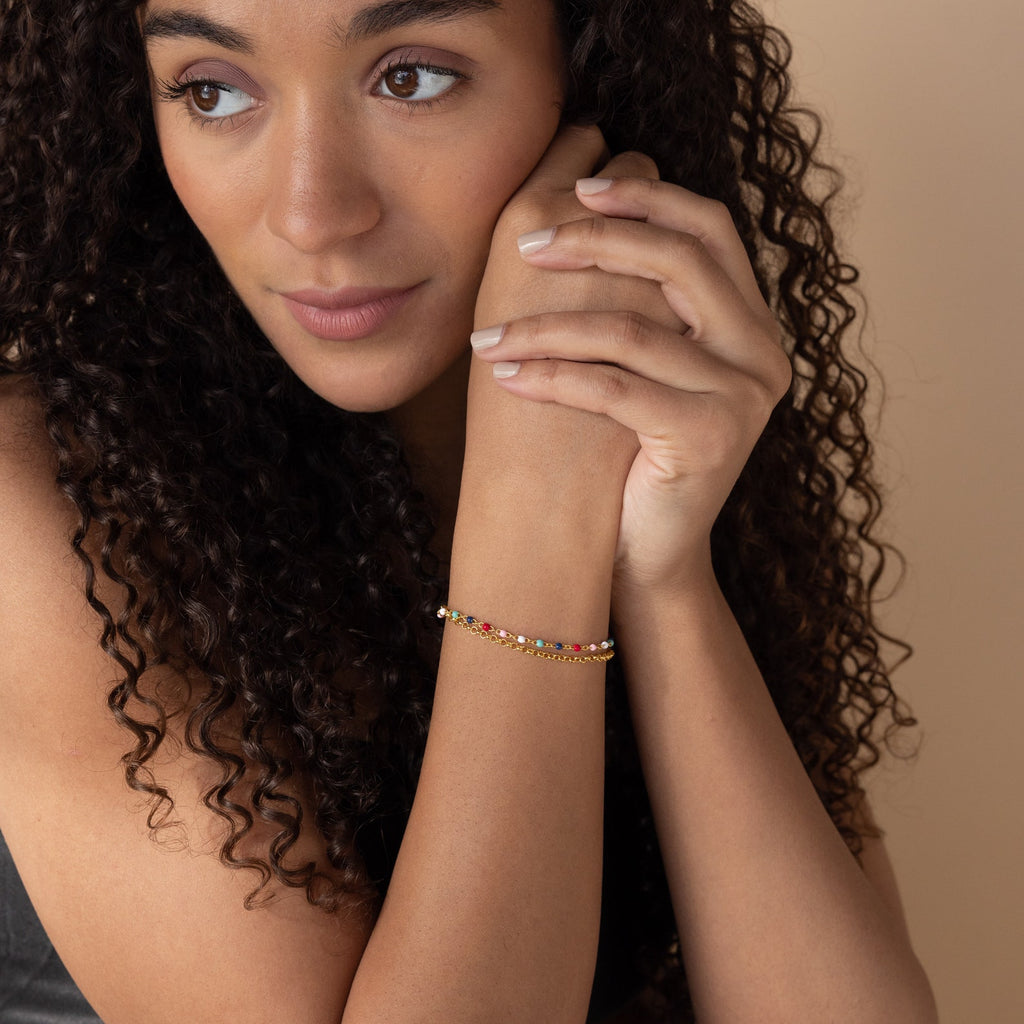 A woman with curly hair wears the Rainbow Station Bracelet, resting her chin on her hands and looking thoughtful.