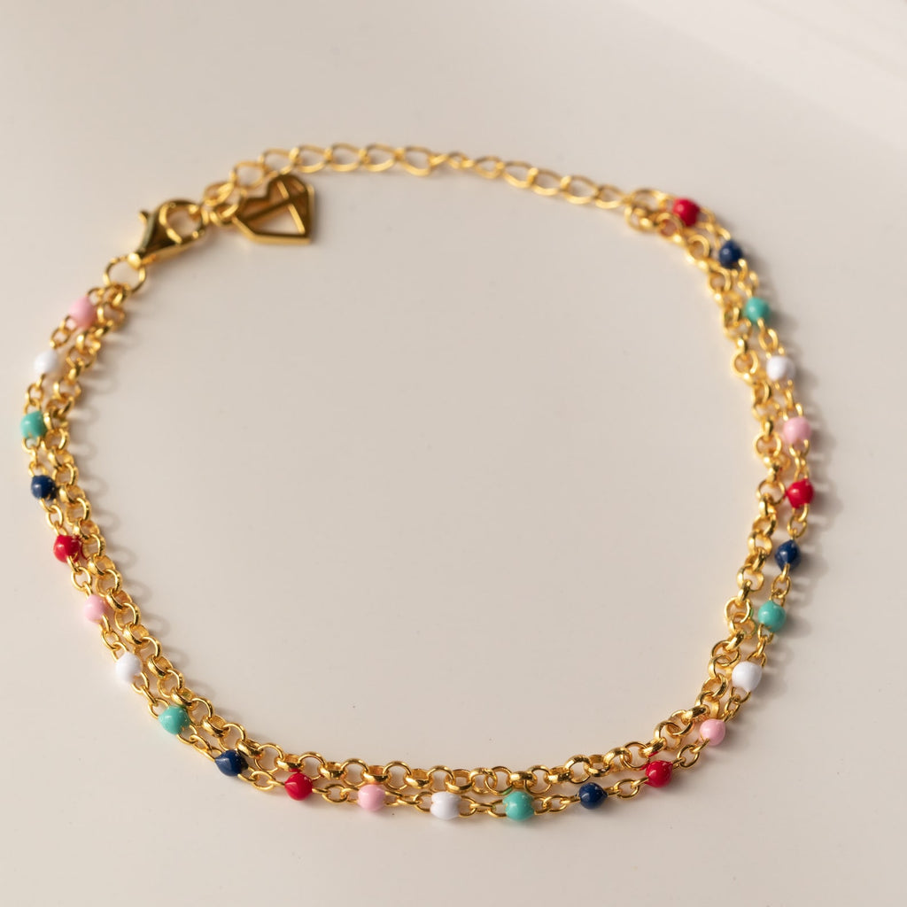 The Rainbow Station Bracelet is a handmade piece featuring a gold double chain with colorful beads and a heart-shaped clasp, displayed on a light surface.
