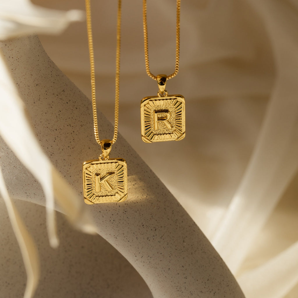 Two Initial Rectangle Pendant Necklaces in gold, each engraved with the letters K and R, are displayed on a neutral background.