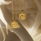 Two Initial Rectangle Pendant Necklaces in gold, each engraved with the letters K and R, are displayed on a neutral background.