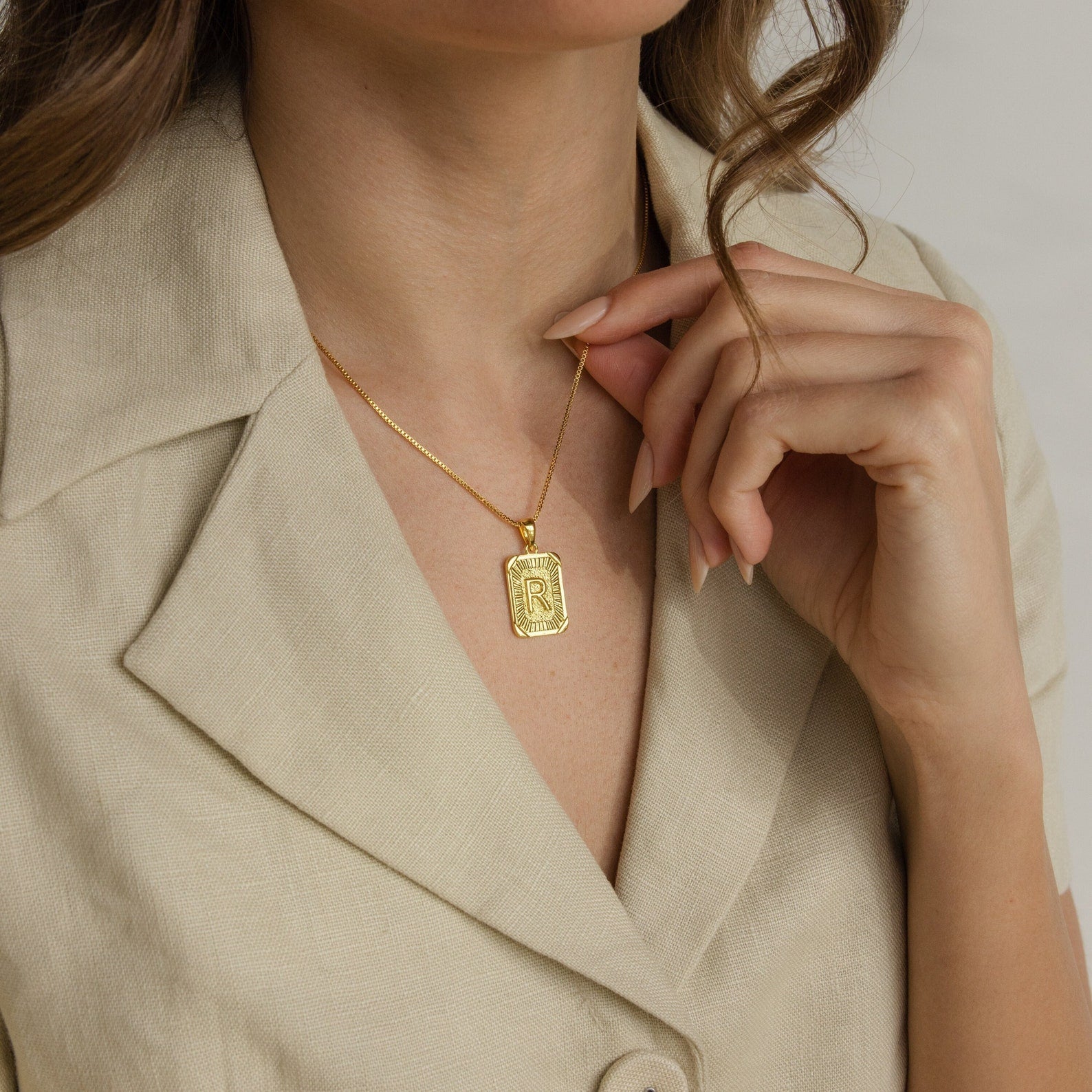 A woman wears a beige blazer and displays the Initial Rectangle Pendant Necklace, featuring a gold rectangle pendant engraved with the letter R.