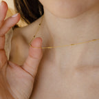 A person gently holds the Bar Station Necklace against their neck, highlighting the subtle beauty of its delicate gold chains.