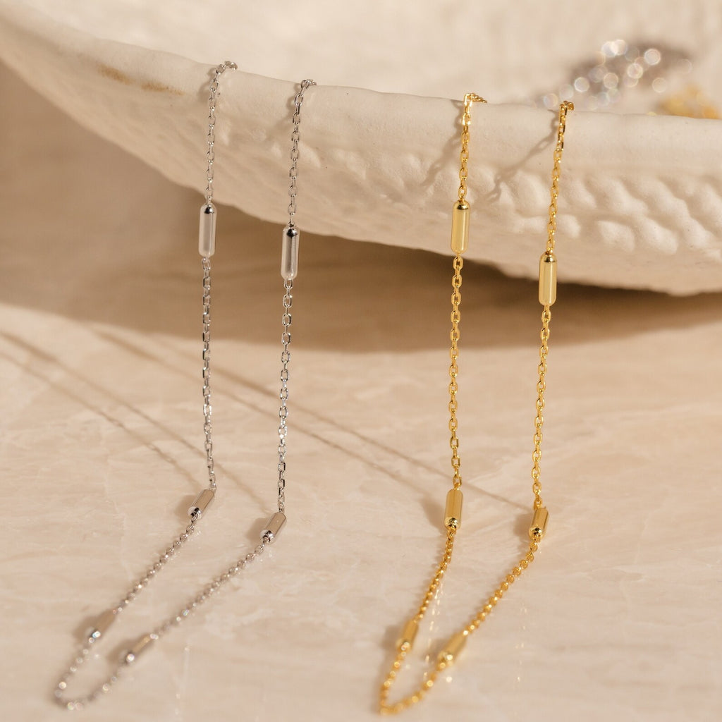 The Bar Station Necklace, featuring one silver and one gold chain each with dainty charms, hangs gracefully over a textured white bowl for an elegant display.