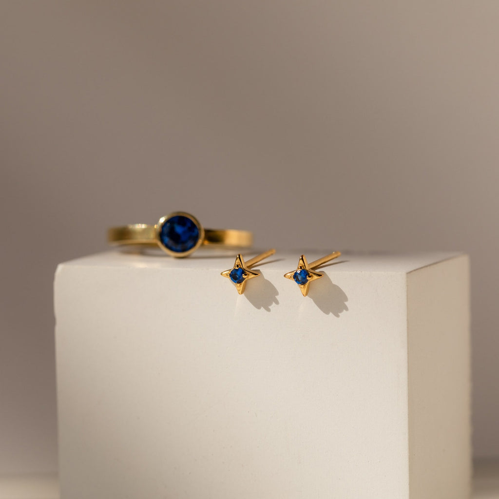 Sapphire Star Studs and a gold ring with a blue stone are elegantly displayed on a white block.